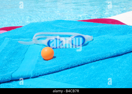 Schutzbrille und orange Kugel auf blaues Handtuch am Pool. Stockfoto