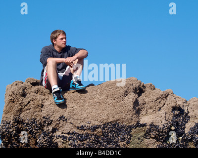 Teenager Junge saß auf einem Muschel bedeckt Felsen, unterstützt von einem klaren blauen Himmel Stockfoto