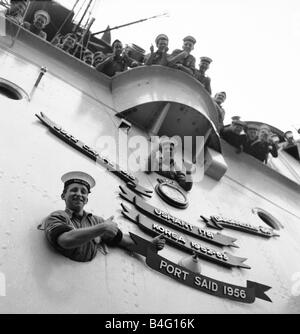 Suez Krise 1956 die Flugzeugträger HMS Ocean verleiht Port Said der Rolle des Battle Honours angezeigt auf seine Flugdeck, die das Schiff nur in Devonport angedockt hat, nach der Rückkehr aus Suez gelehnt aus den Bullaugen, ein Daumen hoch Junior Seemann John Higgins 17 von Oldham unten und Junior Seemann Len Ball 17 von Manchester Stockfoto