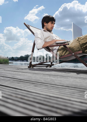 Ein junger Mann sitzt in einem Liegestuhl auf einem Steg mit einem laptop Stockfoto
