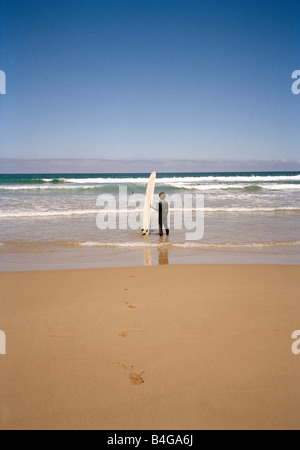 Ein Surfer stehen am Strand mit seinem Surfbrett im Ozean beobachten Stockfoto