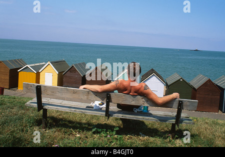 Frau sitzt auf Holzbank auf grasbewachsenen Felsen über hölzerne Strandhütten direkt am Meer in Herne Bay in Kent Stockfoto