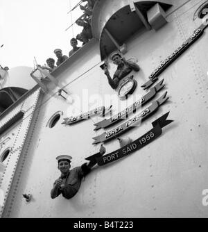 Suez Krise 1956 die Flugzeugträger HMS Ocean verleiht Port Said der Rolle des Battle Honours angezeigt auf seine Flugdeck, die das Schiff nur in Devonport angedockt hat, nach der Rückkehr aus Suez gelehnt aus den Bullaugen, ein Daumen hoch Junior Seemann John Higgins 17 von Oldham unten und Junior Seemann Len Ball 17 von Manchester Stockfoto