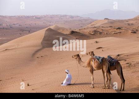 Oman, Sharqiyah, Wahiba Sands. Eine Beduinen kniet in der Wüste beten hält seinen Kamelen durch ihre Halfter zu verhindern, dass sie ins Blaue hinein unter DunesOman OM3 John Warburton-Lee Stockfoto