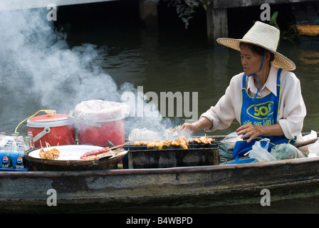 Ältere Frau Grillen von Fleisch auf einem kleinen Grill in einem traditionellen Holzboot auf einem Kanal in Thailand Stockfoto