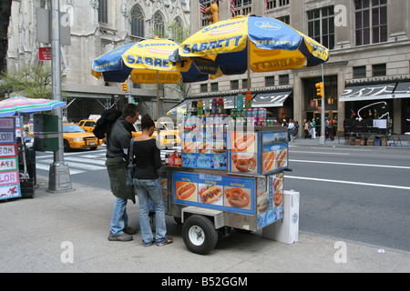 Ein Touristenpaar, das Artikel von einem Lebensmittelhändler der Fifth Ave kauft, mit einem legendären New York City Hot Dog Cart, der mit Sabrett-Schirmen geschmückt ist. Stockfoto