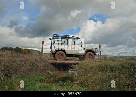 Weiße Land Rover Defender 90, ausgestattet mit einer externen Überrollkäfig, einen kleinen Fluss über eine Holzbrücke überqueren gemacht aus Baumstämmen. Stockfoto