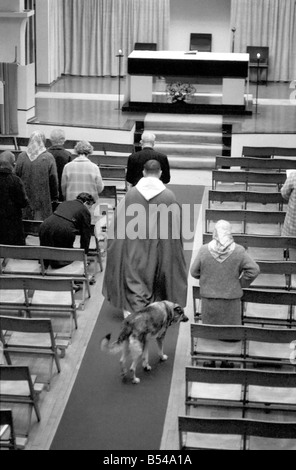 Hunde Tiere humor Religion: "Schatten" der elsässischen ihrem Meister - Pfarrer Dom Edmund Jones - überall er geht folgt, selbst wenn er die Messe in der Kirche Christkönig, Cockfosters, sagt Schatten ist direkt hinter ihm. Sie folgt ihrem Meister in der Kirche und liegt ruhig am Fuße der Wallfahrtskirche Schritte unter dem Altar und befindet sich direkt hinter ihm, wenn der Dienst beendet ist. Dom Edmund hat sogar Sonderwünsche, dass der Hund Hochzeitsservice eingeladen werden, und die Kinder gehen in die Kirche, den Hund zu sehen. Nach seinem master Dom Edmund Jones entlang den kirchlichen Gang Schatten Stockfoto