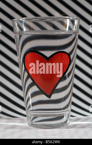 Glas Wasser mit einem roten Herz gegen Diagional schwarzen und weißen Streifen Stockfoto
