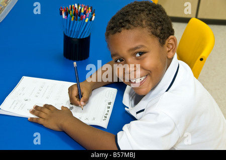 Ein Junge aus der Schule, der Afro-American-Black lernt, im Kindergarten-Klassenzimmer am Schreibtisch zu schreiben Stockfoto