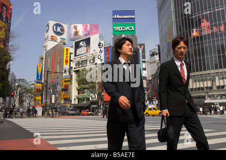 Gut gekleidete junge Männer zu Fuß auf Zebrastreifen mit Werbeplakaten und riesig, Shibuya, Tokyo, Japan Stockfoto