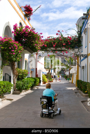 Ältere behinderte Frau im Urlaub mit ihrem motorisierten Roller im Rollstuhl freundlich Fußgängerweg Puerto Mogan Gran Canaria Stockfoto