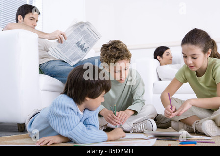 Drei Kinder sitzen auf dem Boden, Färbung, Eltern im Hintergrund lesen Stockfoto