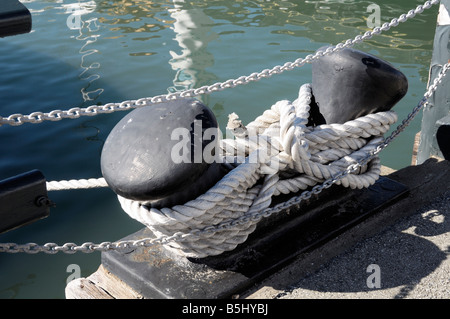 Ein dickes weißes Seil ist in einem sicheren Knoten um einen schwarzen doppelköpfigen Poller auf einem Betondock neben Wasser- und Metallketten gebunden. Stockfoto