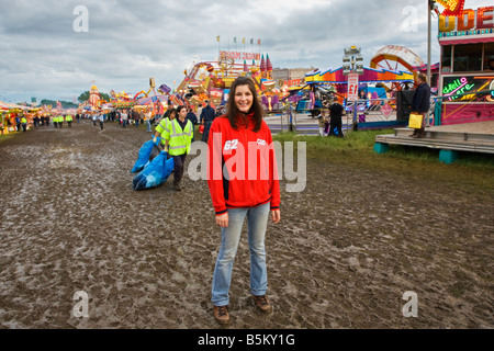 Eine junge Frau posiert für ein Foto vor verschiedenen Fahrten auf dem Jahrmarkt in einem schlammigen Feld Stockfoto
