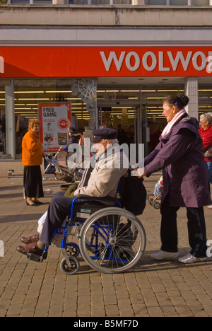 Frau treibt älteren Mann im Rollstuhl in der geschäftigen britischen High street Stockfoto