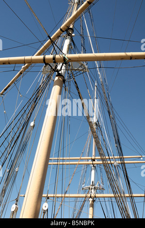 Braune Holzmasten eines Segelschiffes kreuzen sich gegen einen klaren blauen Himmel, durchzogen von einem komplexen Netz aus dunklen Takelinien und Riemenscheiben. Stockfoto