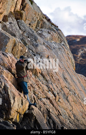 Mann mit Outdoor-Bekleidung Stand auf halber Höhe kleine Felswand Blick auf das Meer mit seinen Händen, die Augen vor der Sonne schützt. Stockfoto