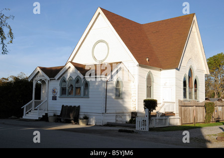 Eine kleine weiße Holzkirche mit einem braunen Giebeldach und Bogenfenstern steht tagsüber entlang einer asphaltierten Straße unter klarem Himmel. Stockfoto