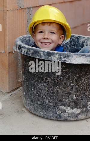 Lächelnde Knabe trug einen gelben Schutzhelm sitzen in einem Gips-Mörtel-Eimer auf einer Baustelle Haus Stockfoto