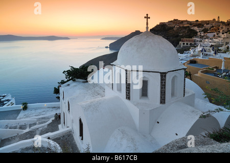 Weiße Kuppel-Kirche mit einem Kreuz bei Sonnenuntergang vor dem Meer und der vulkanischen Insel von Nea Kameni, Santorin, Kykladen, Griechenland Stockfoto