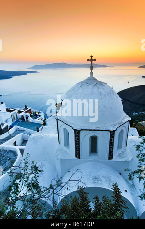 Weiße Kuppel-Kirche mit einem Kreuz bei Sonnenuntergang vor dem Meer und der vulkanischen Insel von Nea Kameni, Santorin, Kykladen, Griechenland Stockfoto