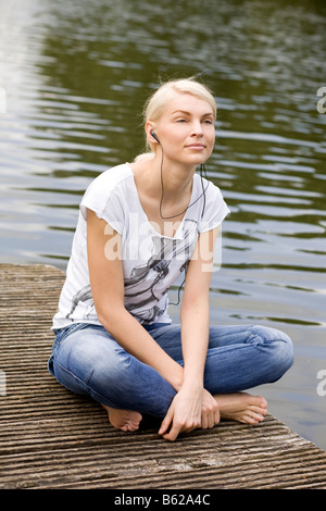 Junge blonde Frau auf einer Werft an einem See zu sitzen und Musik hören Stockfoto