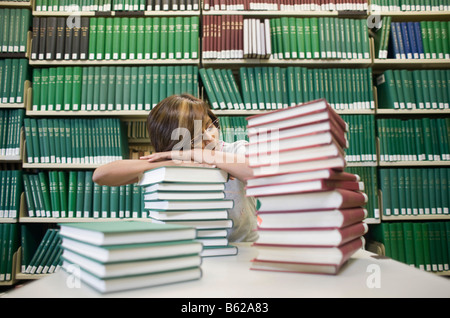 Junge dunkelhaarige Studentin sitzt schlafend mit vielen Büchern an einem Tisch vor einem Bücherregal in einer Bibliothek Stockfoto