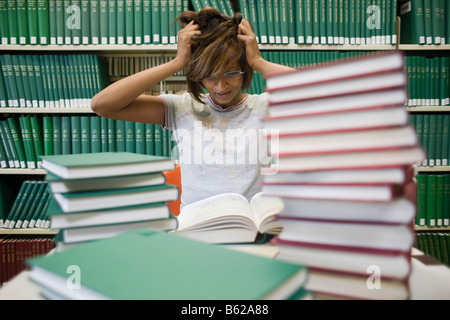 Junge dunkelhaarige Studentin sitzt verzweifelt mit vielen Büchern an einem Tisch vor einem Bücherregal in einer Bibliothek Stockfoto