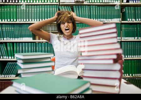 Junge dunkelhaarige Studentin sitzt verzweifelt mit vielen Büchern an einem Tisch vor einem Bücherregal in einer Bibliothek Stockfoto