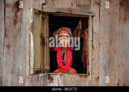 Junge Frau des Stammes Akha Pala Blick aus einem Fenster gekleidet in einem bunten Kopf-Kleid und eine rote Halskette, Ban Saenkham T Stockfoto