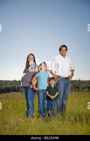 Vierköpfige Familie, einschließlich eine werdende Mutter, zusammen in einem Feld stehen lächelnd und in die Kamera schaut. Stockfoto