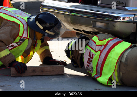 Zwei Feuerwehrleute Holzblöcke unter einem Fahrzeug nach einem Autounfall platzieren, um ihn zu stabilisieren, bevor Sie mit Bergung Stockfoto