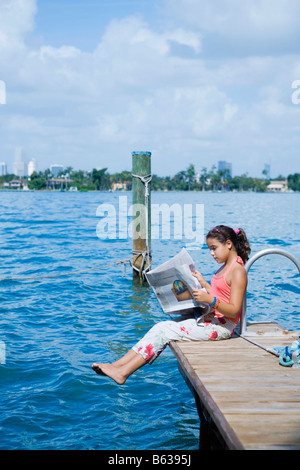 Seitenansicht eines Mädchens auf dem Steg sitzen und eine Zeitung lesen Stockfoto