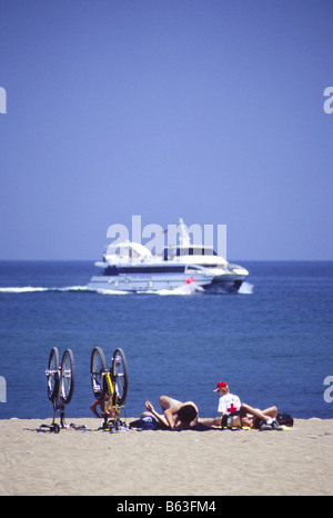 Sonnenbaden am Strand von Barceloneta liegen neben Männer umgedrehten Fahrräder, träge beobachten eines vorbeifahrenden Bootes, Barcelona, Spanien Stockfoto