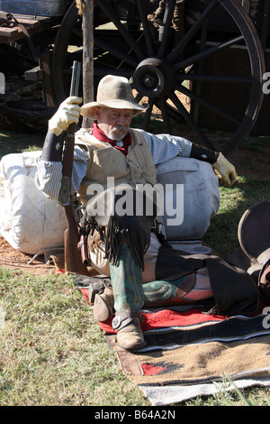 Ein Oldtimer Cowboy lehnte sich gegen seinen Pack und Sattel neben dem Planwagen mit seinem Gewehr Stockfoto