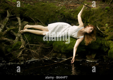 Strawberry blonde Frau in durchscheinenden weißen Baumwoll Unterkleid liegt am Flussufer auf einem Bett aus weichem grünem Moos treiben. Stockfoto