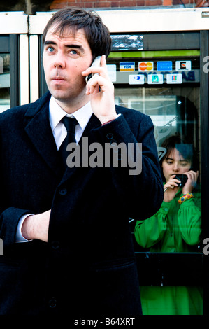 ein Mann mit Hemd und Krawatte auf einem Mobiltelefon im Gespräch mit einer Frau hinter ihm in grünem Kleid in einem Telefongespräch Kasten, UK Stockfoto