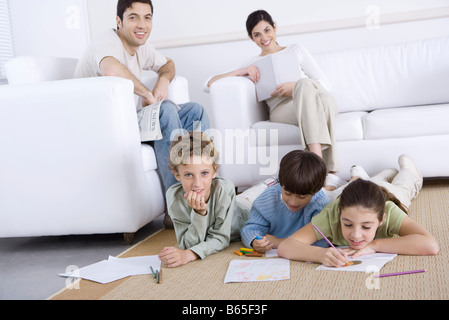 Drei Kinder Färbung auf dem Boden im Wohnzimmer, Eltern entspannend im Hintergrund Stockfoto