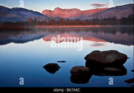 Dawn Licht erhellt die Langdale Pikes mit einem rosigen Glanz spiegelt sich in den stillen Wassern des Elterwater Lake District England UK Stockfoto