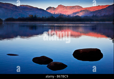 Dawn Licht erhellt die Langdale Pikes mit einem rosigen Glanz spiegelt sich in den stillen Wassern des Elterwater Lake District England UK Stockfoto