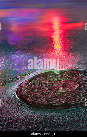 Kanaldeckel in der Nacht mit bunten Neonröhren reflektiert nasse Straße Bürgersteig Stockfoto