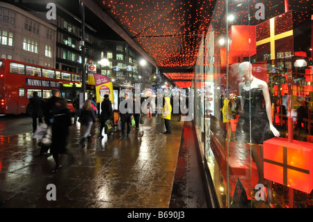 Käufer auf nasser Fahrbahn außerhalb Department Store Fenster vorne mit Weihnachtsgeschenke auf Anzeige der Oxford Street Weihnachtsbeleuchtung Regen in London West End UK Stockfoto