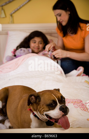 Hispanic Mutter kümmert sich um ihre kranke Tochter im Bett mit dem Familienhund am Fuße des Bettes. Stockfoto