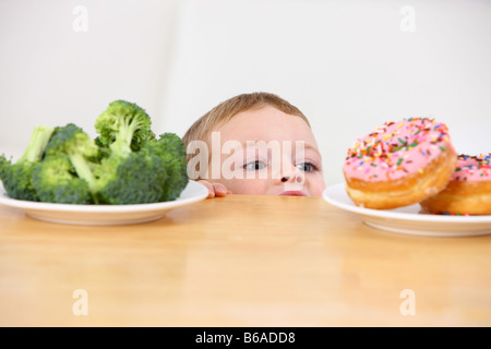 Kleiner Junge peeking über Tabelle auf Platten von Donuts und Brokkoli Stockfoto