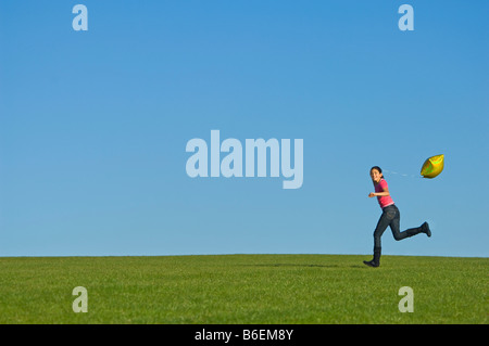 Ein junges Mädchen (11) läuft glücklich über grünen Rasen mit einem gelben Ballon hinter und blauer Himmel. Stockfoto