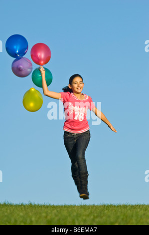 Ein junges Mädchen (11) läuft glücklich über grünen Rasen mit einer Reihe von bunten Luftballons hinter und blauer Himmel. Stockfoto