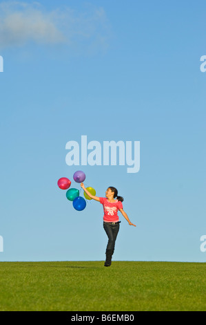 Ein junges Mädchen (11) läuft glücklich über grünen Rasen mit einer Reihe von bunten Luftballons hinter und blauer Himmel. Stockfoto