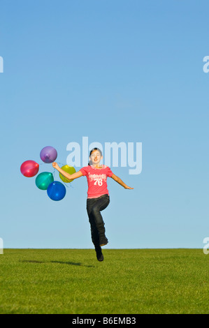 Ein junges Mädchen (11) läuft glücklich über grünen Rasen mit einer Reihe von bunten Luftballons hinter und blauer Himmel. Stockfoto