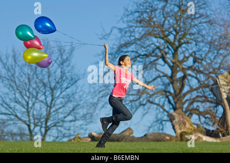 Ein junges Mädchen (11) läuft glücklich über grünen Rasen mit einer Reihe von bunten Luftballons hinter und blauer Himmel. Stockfoto